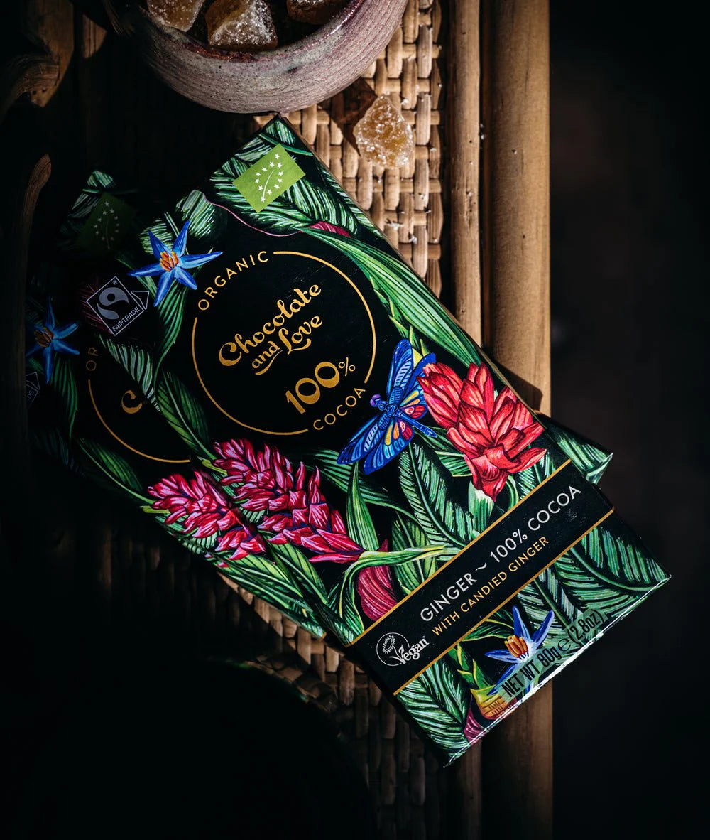Eine Schokoladenverpackung mit einem farbenfrohen Design aus grünen Blättern, roten Blumen und einem blauen Schmetterling. Der Text auf der Verpackung weist darauf hin, dass es sich um ein Bio-Produkt handelt, das zu 100 % aus Kakao besteht und kandierten Ingwer von Chocolate and Love enthält.