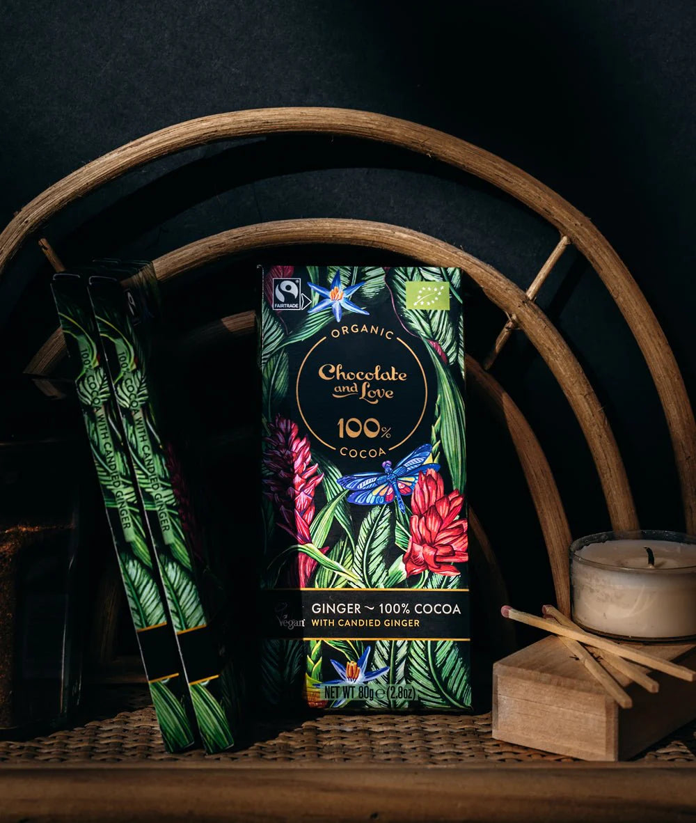 Eine Schokoladenverpackung mit einem farbenfrohen Design aus grünen Blättern, roten Blumen und einem blauen Schmetterling. Der Text auf der Verpackung weist darauf hin, dass es sich um ein Bio-Produkt handelt, das zu 100 % aus Kakao besteht und kandierten Ingwer von Chocolate and Love enthält.