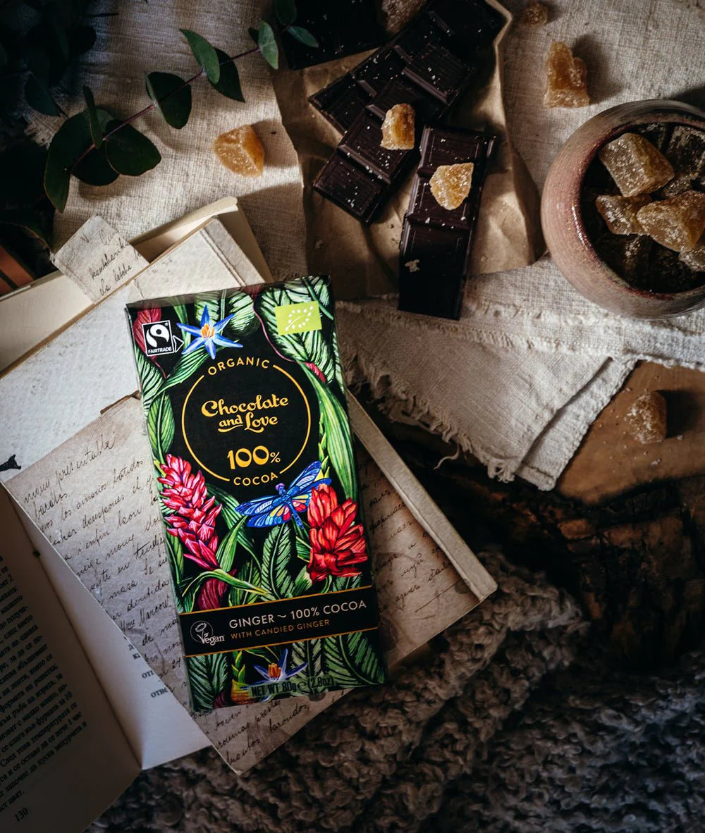 Eine Schokoladenverpackung mit einem farbenfrohen Design aus grünen Blättern, roten Blumen und einem blauen Schmetterling. Der Text auf der Verpackung weist darauf hin, dass es sich um ein Bio-Produkt handelt, das zu 100 % aus Kakao besteht und kandierten Ingwer von Chocolate and Love enthält.